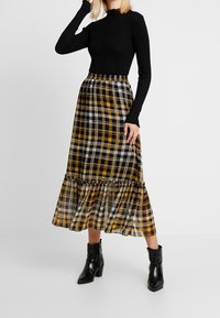 Schwarzes, geripptes Langarmoberteil kombiniert mit einem gelb-schwarz karierten Midikleid mit rüschigem Saum, getragen mit schwarzen Knöchelstiefeln.