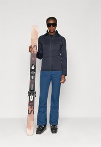 Equipo de esquí: persona vestida con una chaqueta de cremallera azul marino, pantalones azules, botas de esquí negras, sosteniendo un esquí rosa y marrón con fijaciones negras.