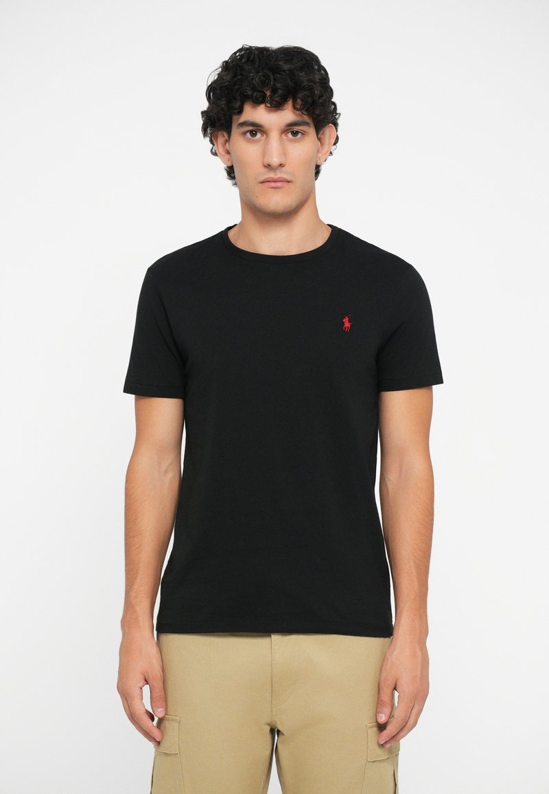 Juodas medvilninis marškinėlis su apvalia kaklo iškirpte, trumpomis rankovėmis ir mažuoju raudonu išsiuvinėtu logotipu kairėje krūtinės pusėje. Sklandus paviršius.