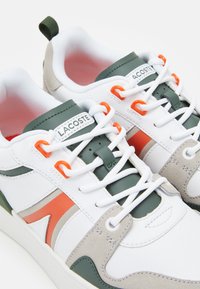 Paire de baskets blanches Lacoste avec des accents verts et orange, des lacets blancs et des détails en daim sur fond blanc.