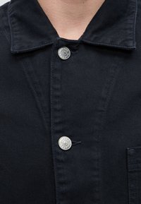 Gros plan du col et de la partie supérieure de la poitrine d'une veste en denim foncé avec des boutons-pression argentés visibles indiquant "SAMSOE SAMSOE."