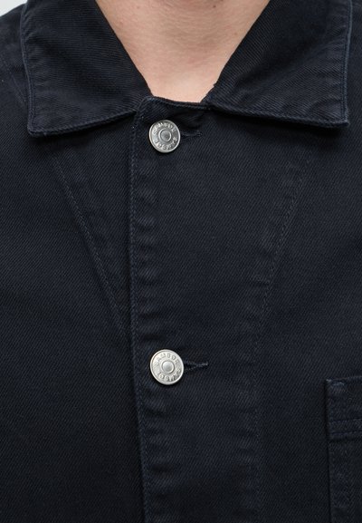 Gros plan du col et de la partie supérieure de la poitrine d'une veste en denim foncé avec des boutons-pression argentés visibles indiquant "SAMSOE SAMSOE."