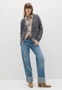Cárdigan gris de punto con botones, camisa de cuadros debajo, jeans de pierna ancha de color azul claro con dobladillos, y botines de tacón color marrón claro.