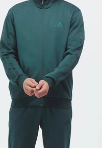 Veste de sport verte foncée avec un col montant, fermeture éclair et accents de logo ton sur ton. Tissu lisse avec des poignets et une taille côtelés.