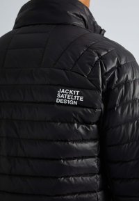 Zwarte pufferjack met een gestikt patroon, voorzien van een hoge kraag en een wit logo "JACK1T SATELITE DES1GN" op de linkerschouder.