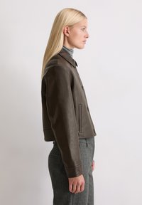 Braune Leder-Cropped-Jacke mit Kragen, einreihigem Design, Seitentaschen und Knopfdetails, kombiniert mit grauen strukturierten Hosen.