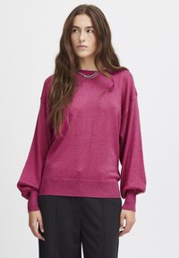 Magenta oversized tröja gjord av ett mjukt, skimrande tyg, med ribbat nederkant och ballongsärmar, ihop med svarta byxor.