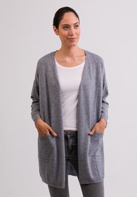 Grijze gebreide cardigan met een losse pasvorm, voorzien van lange mouwen, voorzakken en een V-hals ontwerp, gedragen over een wit shirt.