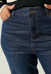 Tummansiniset denim-farkut, joissa on kapea malli. Farkuissa on kultaiset tikkaukset, nappisuljenta ja sivutaskut. Kangas on hieman tekstuuriin.