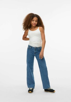 White tank top, blue wide-leg jeans, and black sneakers with light soles. Model poses with one hand on hip, displaying natural curly hair.