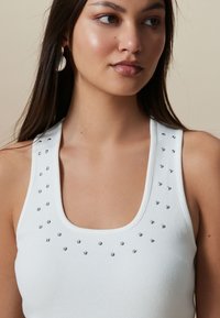 Débardeur blanc à côtes avec un encolure dégagée et des clous argentés soulignant les épaules. Texture lisse et coupe ajustée.