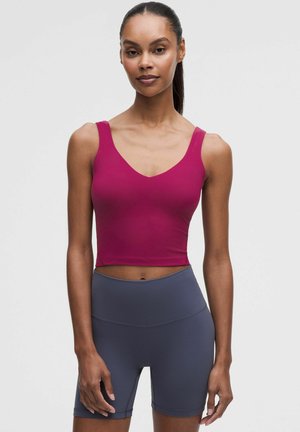 Kvinde iført en tætsiddende ærmeløs magentarød croptop og højtaljede mørkegrå cykelshorts, der står mod en ensfarvet lys baggrund.