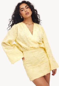 Robe mini jaune avec une finition texturée et effilochée ; elle présente des manches longues et évasées ainsi qu'un décolleté en V profond pour une silhouette moderne.