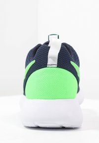 Sportovní bota v tmavě modré a neonově zelené barvě s síťovaným svrškem, bílou podrážkou a poutkem na snadné nazouvání. Vyznačuje se hladkou texturou a kontrastními barvami.