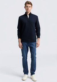 Sudadera azul marino con cierre de cremallera, confeccionada en tejido texturizado, combinada con vaqueros azul claro y zapatillas blancas. Presenta cuello y puños acanalados.