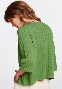 Suéter verde de punto con un patrón de encaje, mangas anchas, dobladillo en forma de onda y un ajuste relajado. La textura es ligera y transpirable.