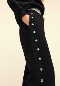 Schwarze Jogginghosen aus weichem Stoff, mit einer Seitentasche und silbernen Druckknöpfen entlang der Außennähte. Elastischer Bund.