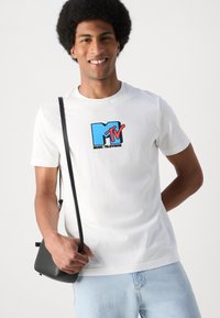 Camiseta de algodón blanca con un prominente logo de MTV en azul y rojo. El modelo sostiene un pequeño bolso negro cruzado y lleva puesto un par de jeans de mezclilla azul claro.
