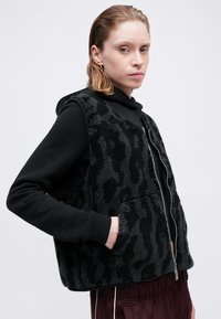 Prsluk s uzorkom leoparda u crnoj i sivoj boji izrađen od teksturiranog flisa. Ima zatvarač sprijeda, dva bočna džepa i opušteni kroj.