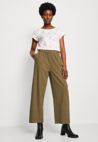 T-shirt blanc avec un motif floral violet et rose, associé à un pantalon wide-leg olive à taille haute. Des bottines noires viennent compléter le look.