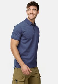 Glimlachende jonge man die een blauw poloshirt en kaki cargobroek draagt, staand en iets naar rechts kijkend tegen een witte achtergrond.