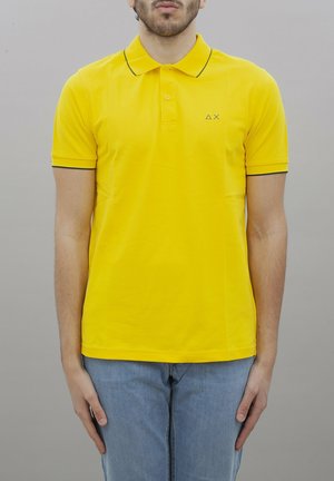 Polo giallo con maniche corte, colletto e bordi neri a contrasto, dotato di un piccolo logo sul lato sinistro del petto. Materiale in cotone.