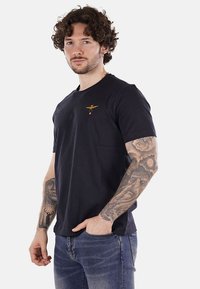 T-shirt corto blu navy in cotone con collo rotondo, caratterizzato da un piccolo emblema d'aquila dorata sul petto e una vestibilità rilassata.