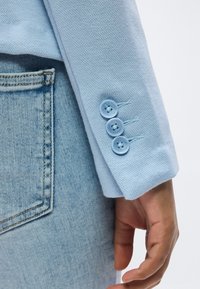 Manche de blazer bleu clair avec trois boutons assortis reposant près de la poche arrière d'un jean en denim délavé clair.