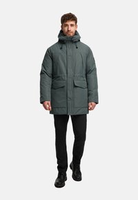 Vadderad parkas i mörkgrön färg med huva, två framfickor och rak skärning. Bärs med svarta byxor och robusta svarta stövlar.