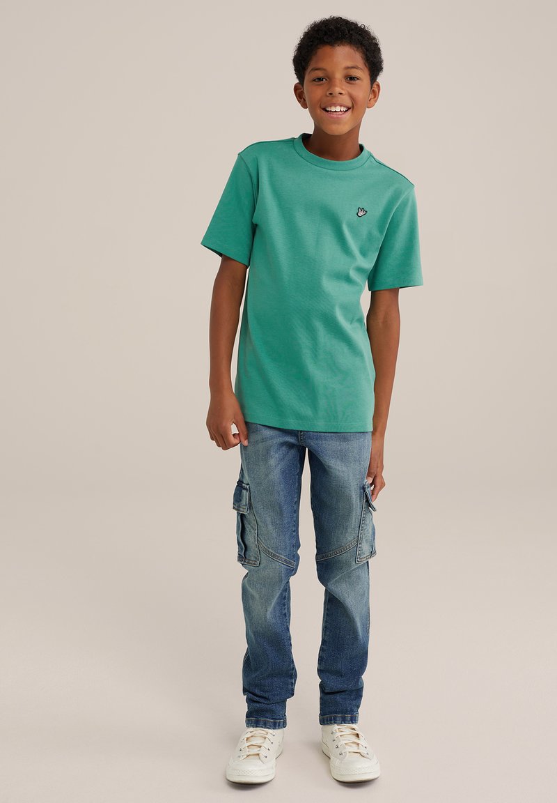 Žalia medvilninė marškinė su trumpomis rankovėmis ir mažai logotipu krūtinėje, derinama su mėlynomis džinsinėmis kargo kelnėmis su šoninėmis kišenėmis.