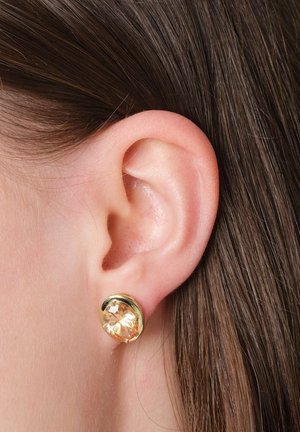 Close-up of an ear with brown hair wearing a round gold earring featuring a large amber-colored gemstone.