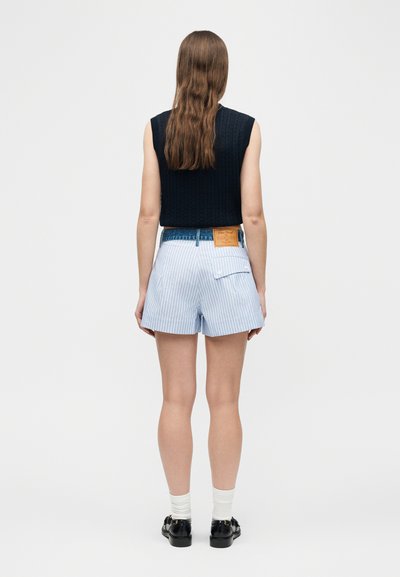 Γυναίκα με μακριά καστανά μαλλιά που φοράει αμάνικο μαύρο τοπ, μπλε και λευκές ριγέ σορτς με πίσω τσέπη, λευκές κάλτσες και μαύρα παπούτσια.