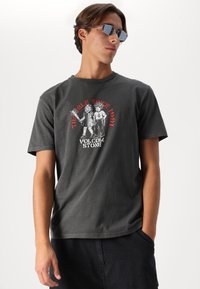 Grijze katoenen t-shirt met een graphic print van twee figuren en de tekst "TRIPLE SINCE 1991" en "VOLCOM STONE" in wit en rood.
