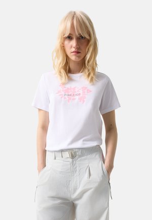 Blondynka w białej koszulce "FIRE + ICE" i jasnych spodniach z paskiem, stojąca z rękami w kieszeniach na jednolitym tle.