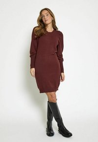 Abito lavorato a maglia bordeaux con maniche lunghe e polsini a coste, caratterizzato da una silhouette aderente, abbinato a stivali al ginocchio in pelle nera.