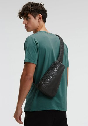 Junger Mann in einem türkisfarbenen Hemd trägt eine schwarze Camp David Umhängetasche auf dem Rücken und schaut seitlich, vor einem einfarbigen Hintergrund.