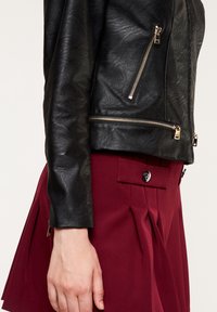 Giacca da motociclista in pelle nera con cerniere dorate, abbinata a una gonna plissettata color burgundy, che mostra una texture liscia e dettagli di sartoria nitidi.