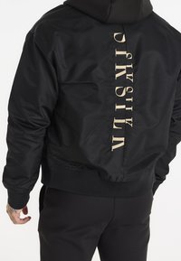Giubbotto bomber nero con finitura lucida, dotato di cappuccio e testo verticale color crema sul retro. Polsini e orlo a costine.