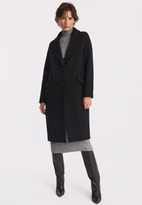 Kobieta w czarnym długim płaszczu na szarej sukience z golfem, nosząca czarne kozaki za kolano, stojąca na tle jednolitej białej ściany.