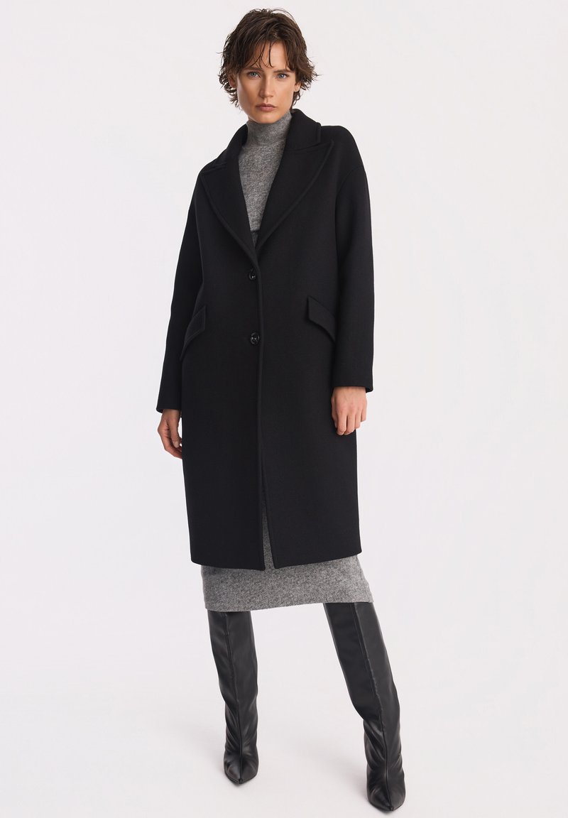 Kobieta w czarnym długim płaszczu na szarej sukience z golfem, nosząca czarne kozaki za kolano, stojąca na tle jednolitej białej ściany.