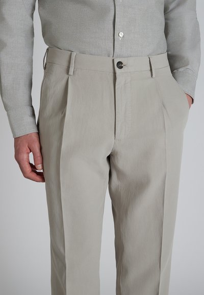 Pantalones beige claros con una textura suave, que presentan dos pliegues frontales, un cierre de botón y bolsillos laterales. Combinados con una camisa gris claro.