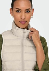 Gilet rembourré beige clair avec un col haut et une fermeture éclair à l'avant, porté sur une chemise vert olive. Présente des sections matelassées horizontales.