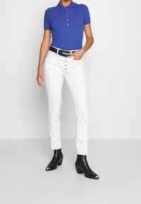 Camisa polo azul con cuello y cinco botones, combinada con jeans rectos blancos que tienen bolsillos frontales y un cinturón negro. Botas negras hasta el tobillo.