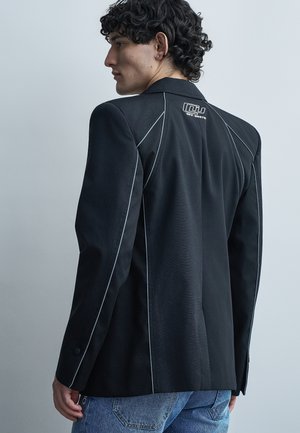 Fiatal férfi göndör hajjal, fekete blézer fehér szegéllyel és logóval a felső háton, kék farmernadrággal, balra nézve.