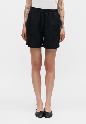 Personne portant un short noir à cordon, un haut blanc sans manches, des chaussures plates noires et des tatouages sur les avant-bras, debout devant un fond uni.