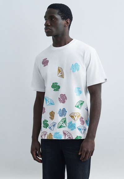 Hombre con camiseta blanca con patrones coloridos de signos de dólar y diamantes, de pie contra un fondo liso de color gris claro.