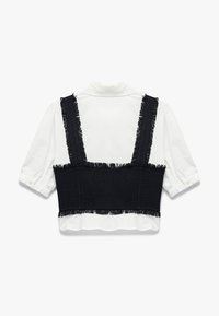 Sort tekstureret crop top med frynset stropper, lagt over en hvid skjorte med puffede ærmer og knaplukninger.