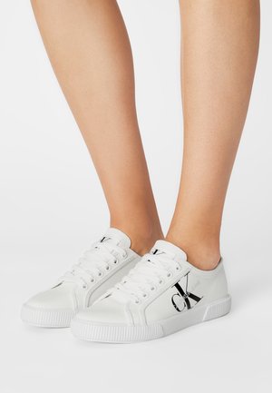 Weiße Leinenschuhe mit geriffelter Sohle, schwarzem Logo und Schnürdesign. Das Obermaterial ist glatt mit minimaler Textur.