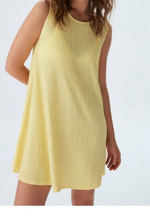 Vrouw in mouwloze, gestructureerde gele mini-jurk staand tegen een effen achtergrond, met één arm gebogen achter haar rug en de andere ontspannen langs haar zij.