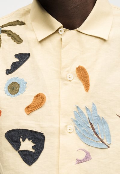 Φωτεινό μπεζ πουκάμισο με κουμπιά και πολύχρωμες αφηρημένες υφασμάτινες εφαρμογές, συμπεριλαμβανομένων μπλε φύλλων και διάφορων σχημάτων στο μπροστινό μέρος.